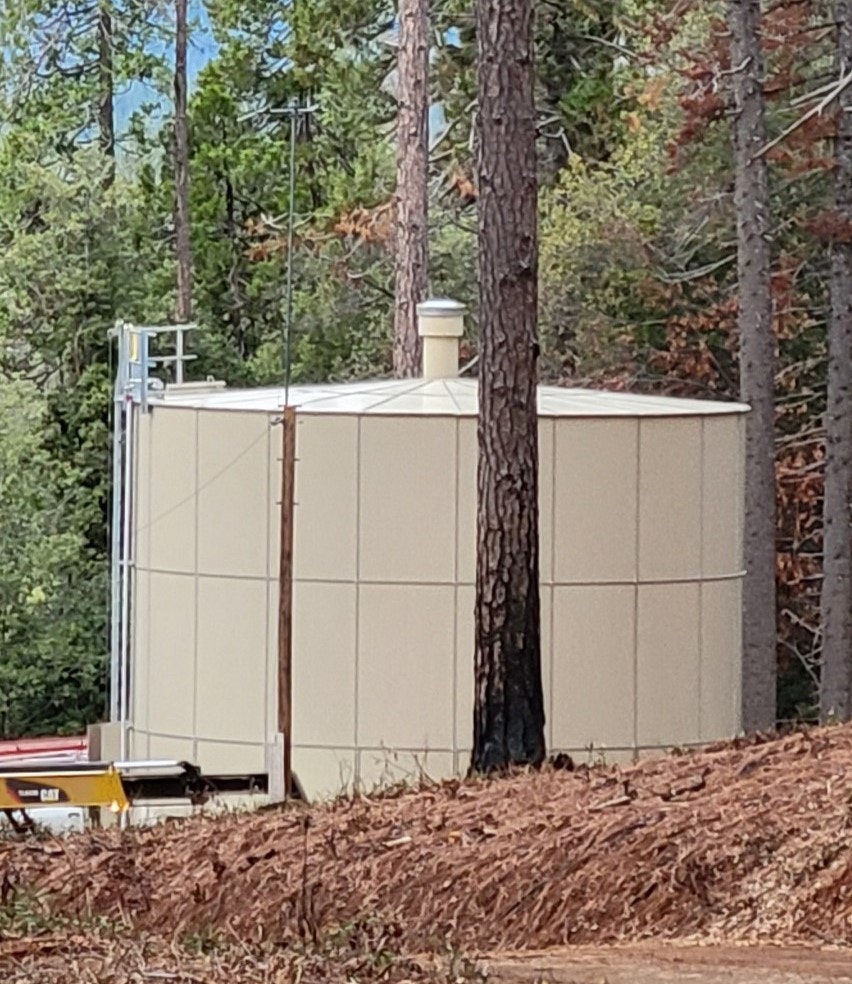 A beige industrial outdoor enclosure among trees with a small antenna and utility equipment nearby.