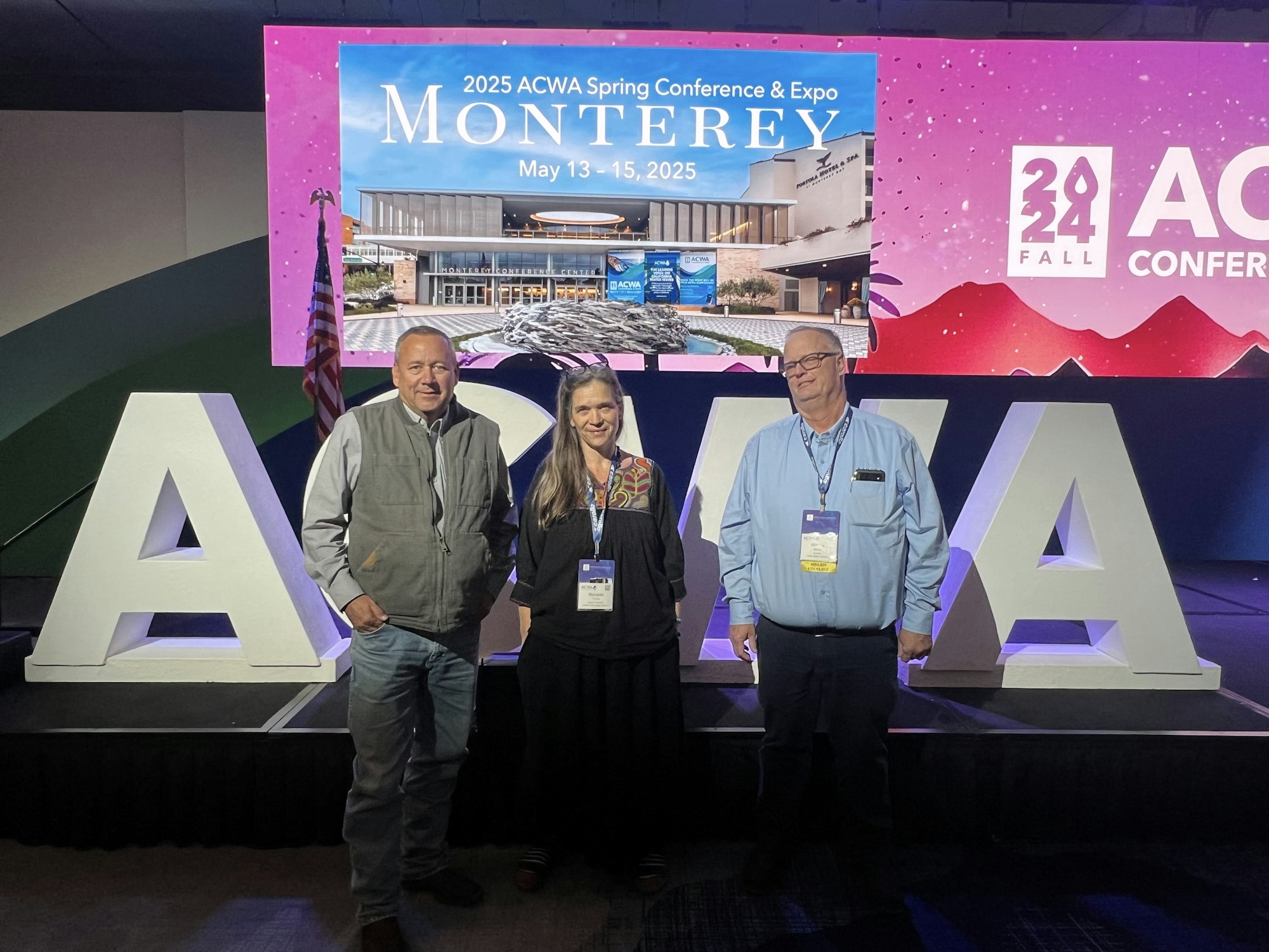 Three people pose in front of a conference display for the 2025 ACWA Spring Conference & Expo in Monterey.