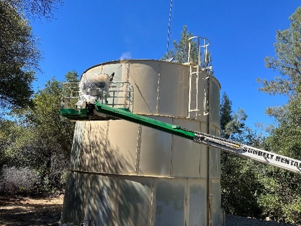 Rackerby Tank with a conveyor belt system against a backdrop of trees and blue sky.