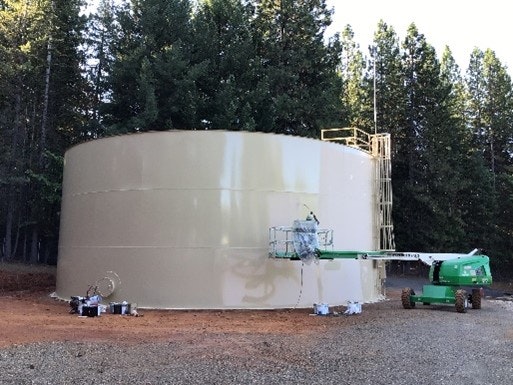 Large storage tank with a person on a boom lift working beside it, surrounded by a forested area.