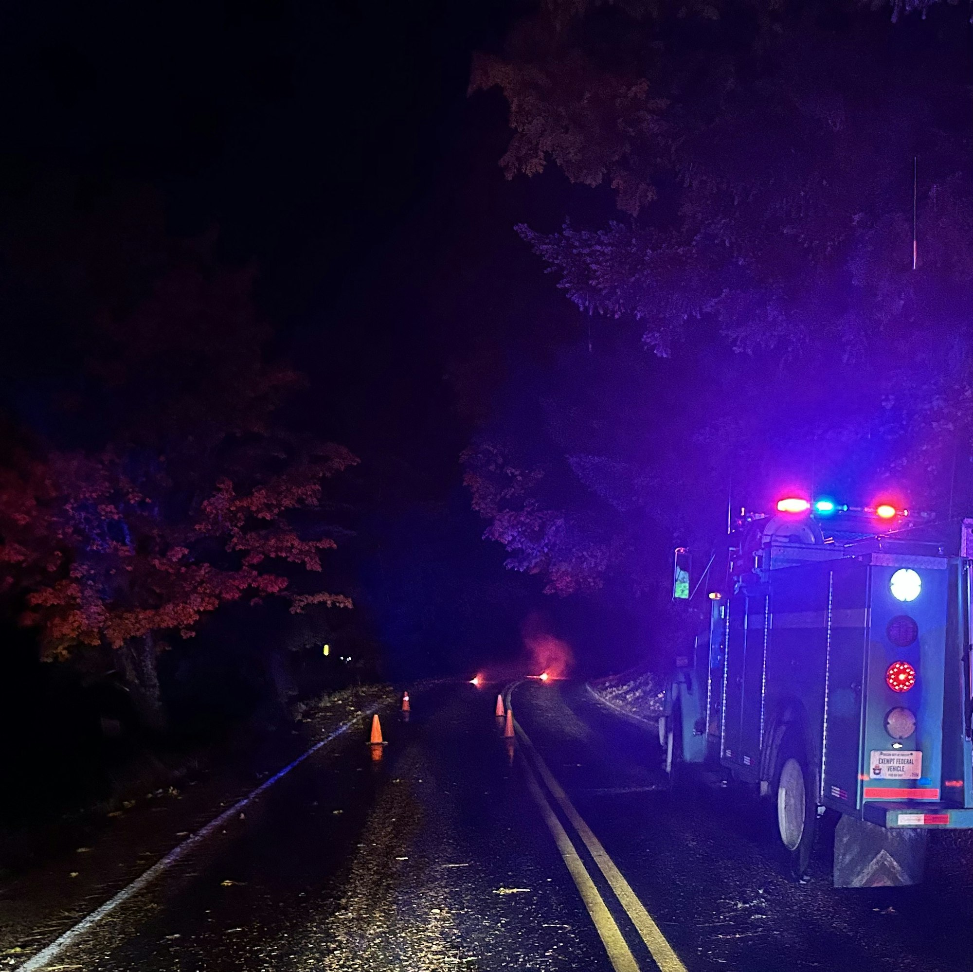 A dark road scene at night with a blue truck, flashing lights, traffic cones, and smoke in the distance.
