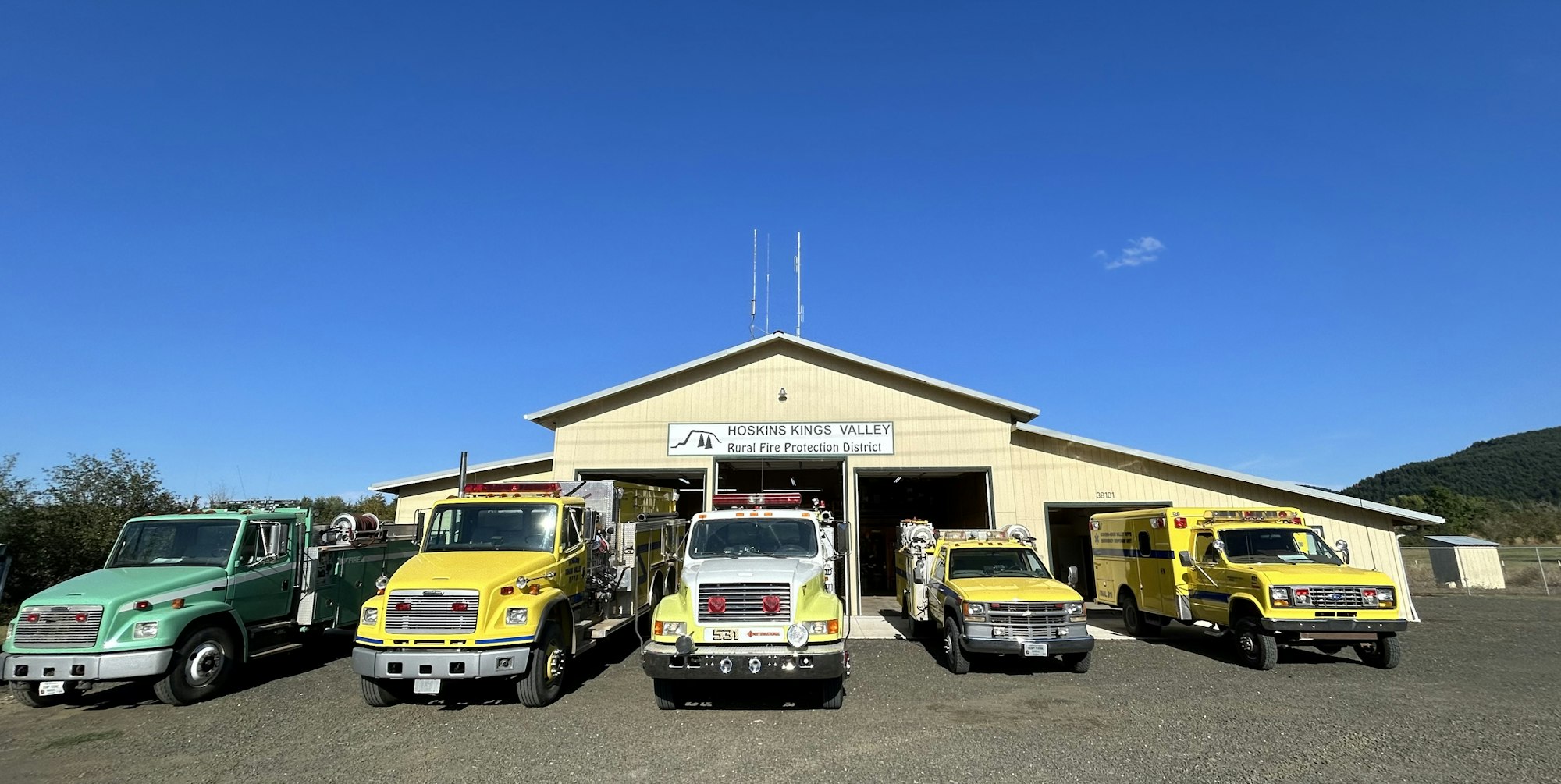 The image shows a fire protection district building with several emergency vehicles parked in front under a clear blue sky.
