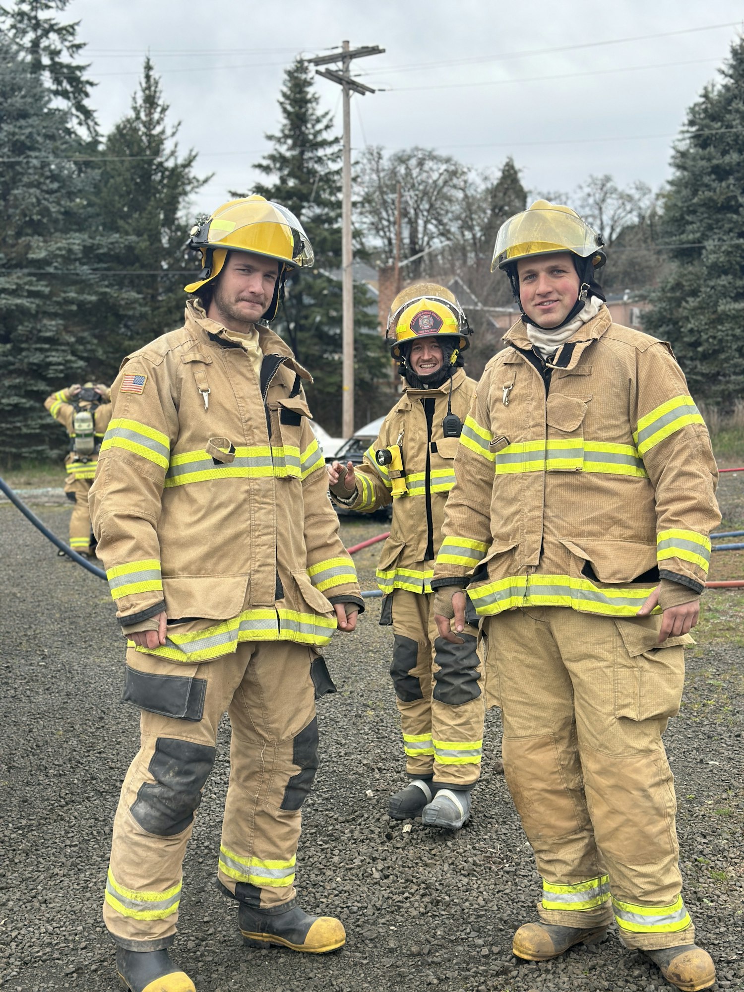 Three firefighters in protective gear pose outdoors, ready for action, with trees and utility poles in the background.