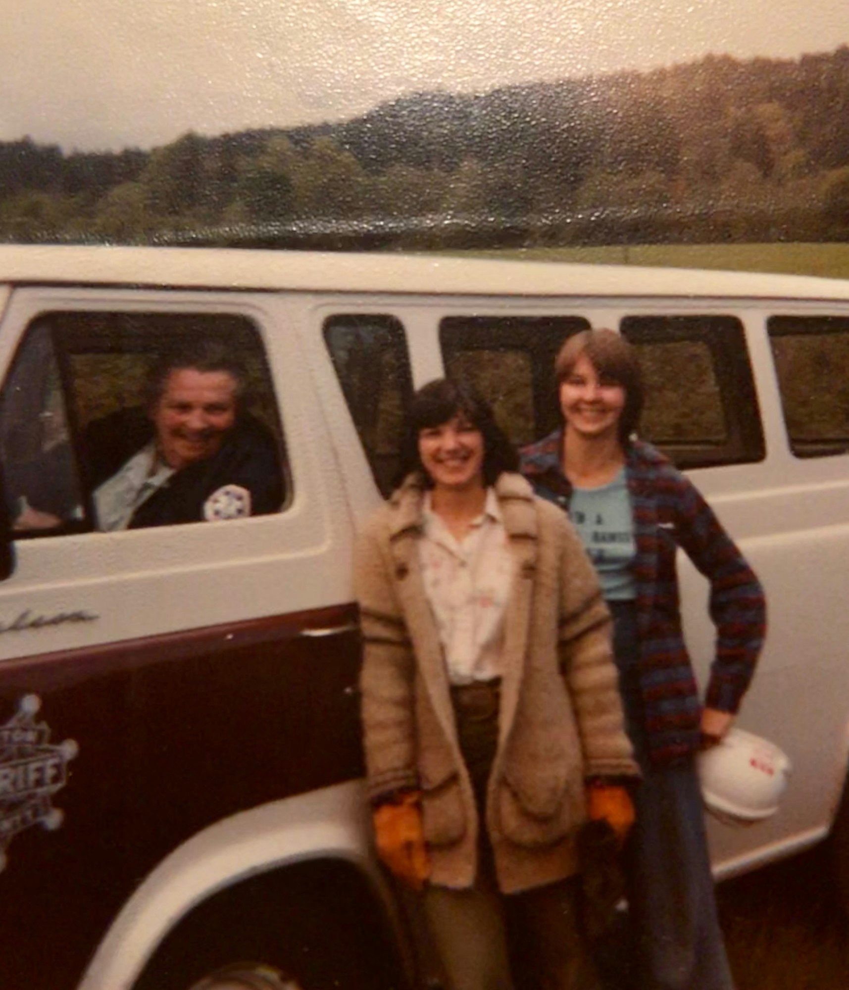 A vintage photo featuring three people near a van, smiling, in a scenic outdoor setting.