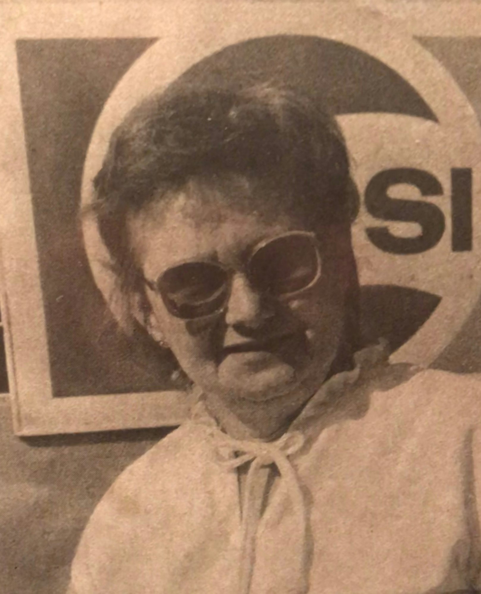 A woman in sunglasses poses, wearing a light-colored outfit, with a sign featuring bold letters in the background.
