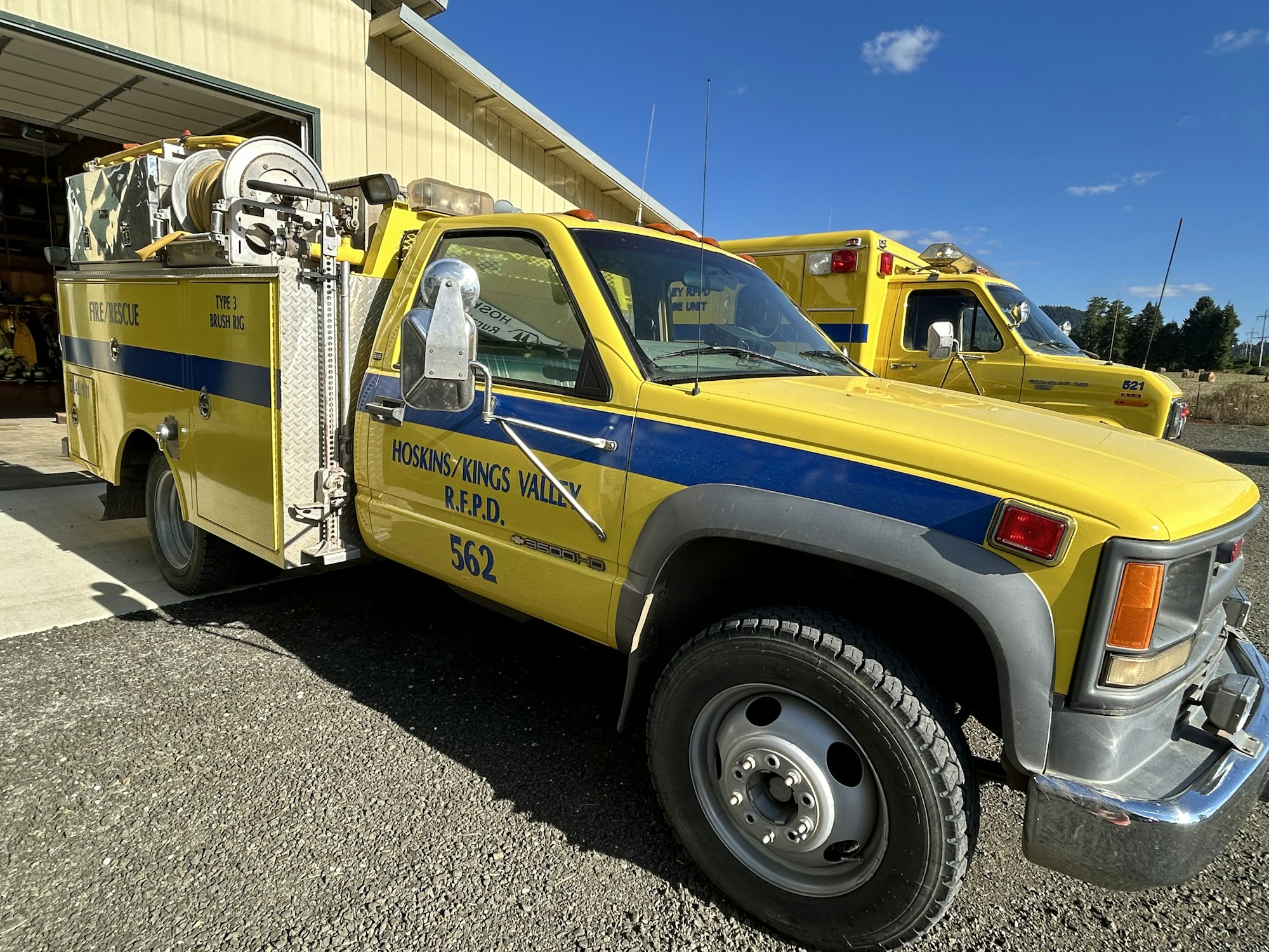 A yellow fire/rescue truck with emergency equipment parked in a garage, alongside another emergency vehicle in the background.