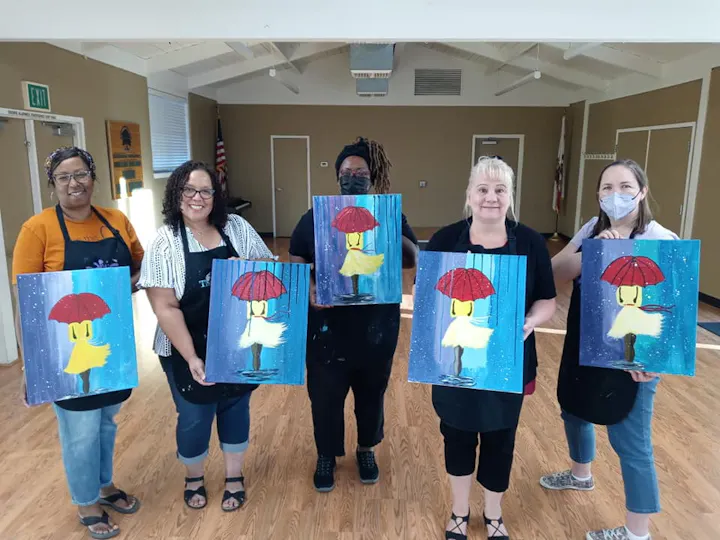 A group of five women proudly hold their paintings of a figure with an umbrella, showcasing their completed artwork together.
