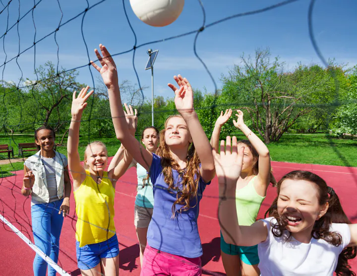 A group of girls is playing volleyball outdoors, joyfully reaching for the ball under a clear blue sky.