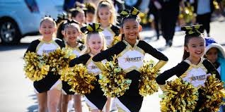 A group of young cheerleaders in black and gold uniforms, holding pom-poms, lined up and smiling during an event.