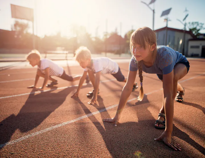 A girl and two boys are in a competitive starting position on a running track, set against a bright, sunny background.