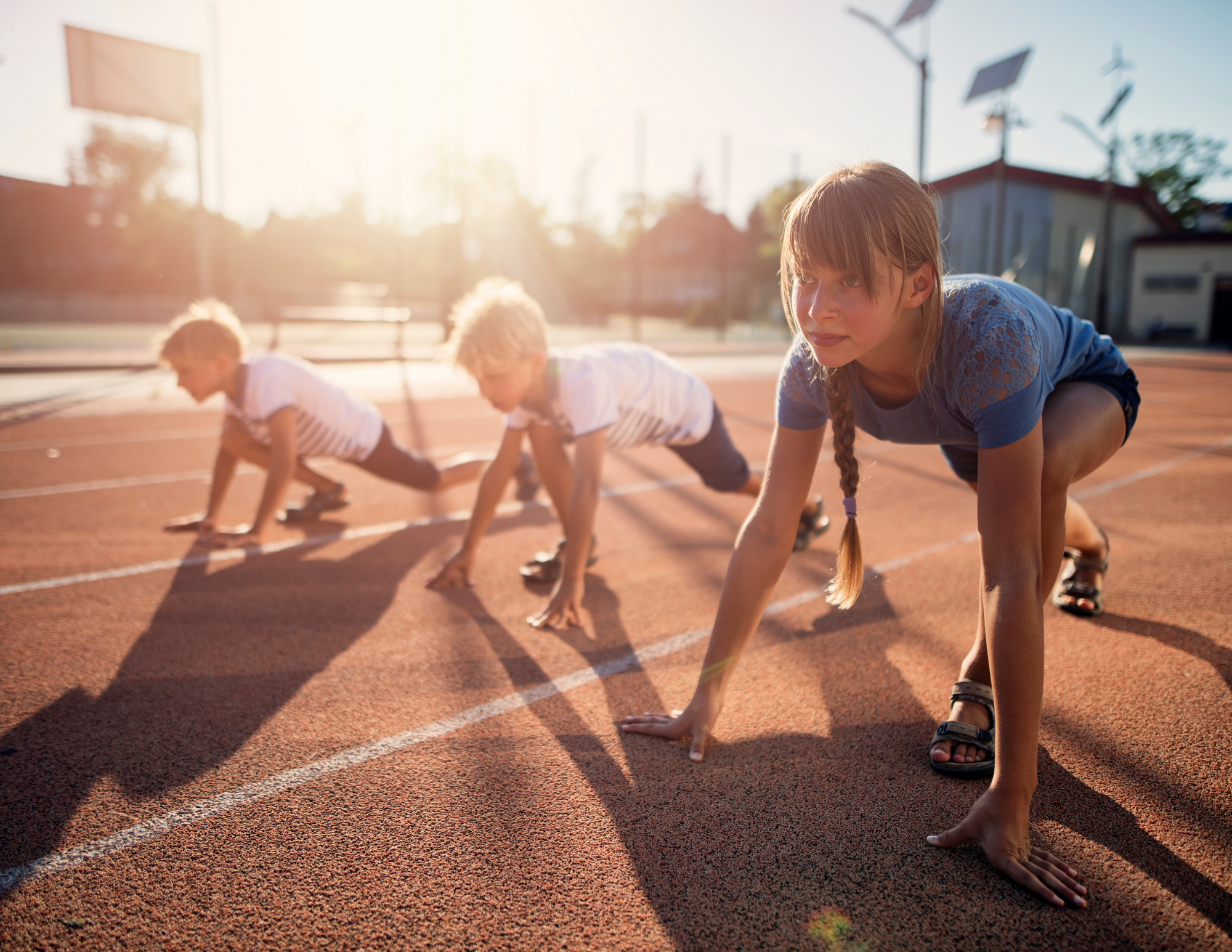 A girl and two boys are in a competitive starting position on a running track, set against a bright, sunny background.