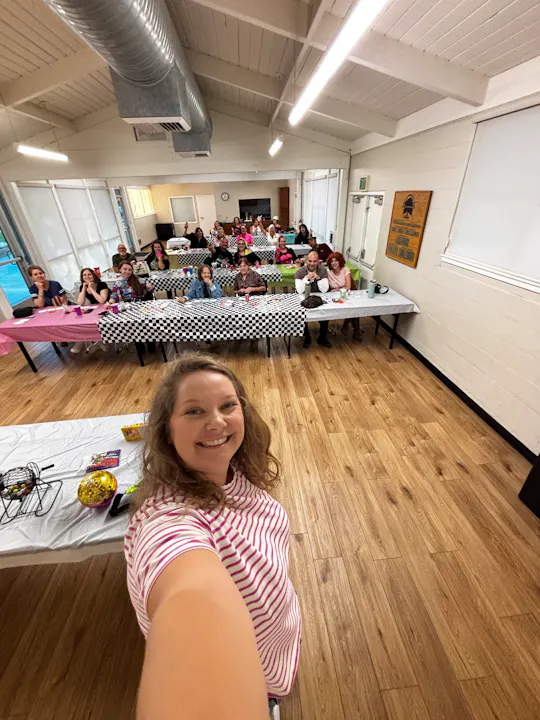A person takes a selfie in a social gathering, featuring a group of people seated at tables with festive decorations.