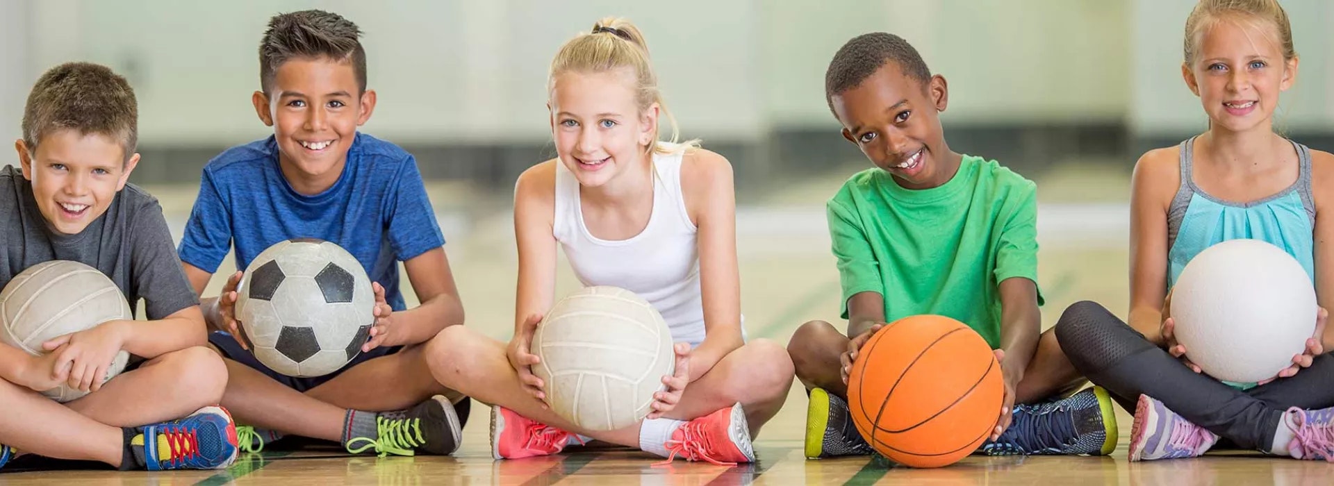 Five kids sit on a court holding different sports balls, smiling and looking at the camera, ready for fun activities.