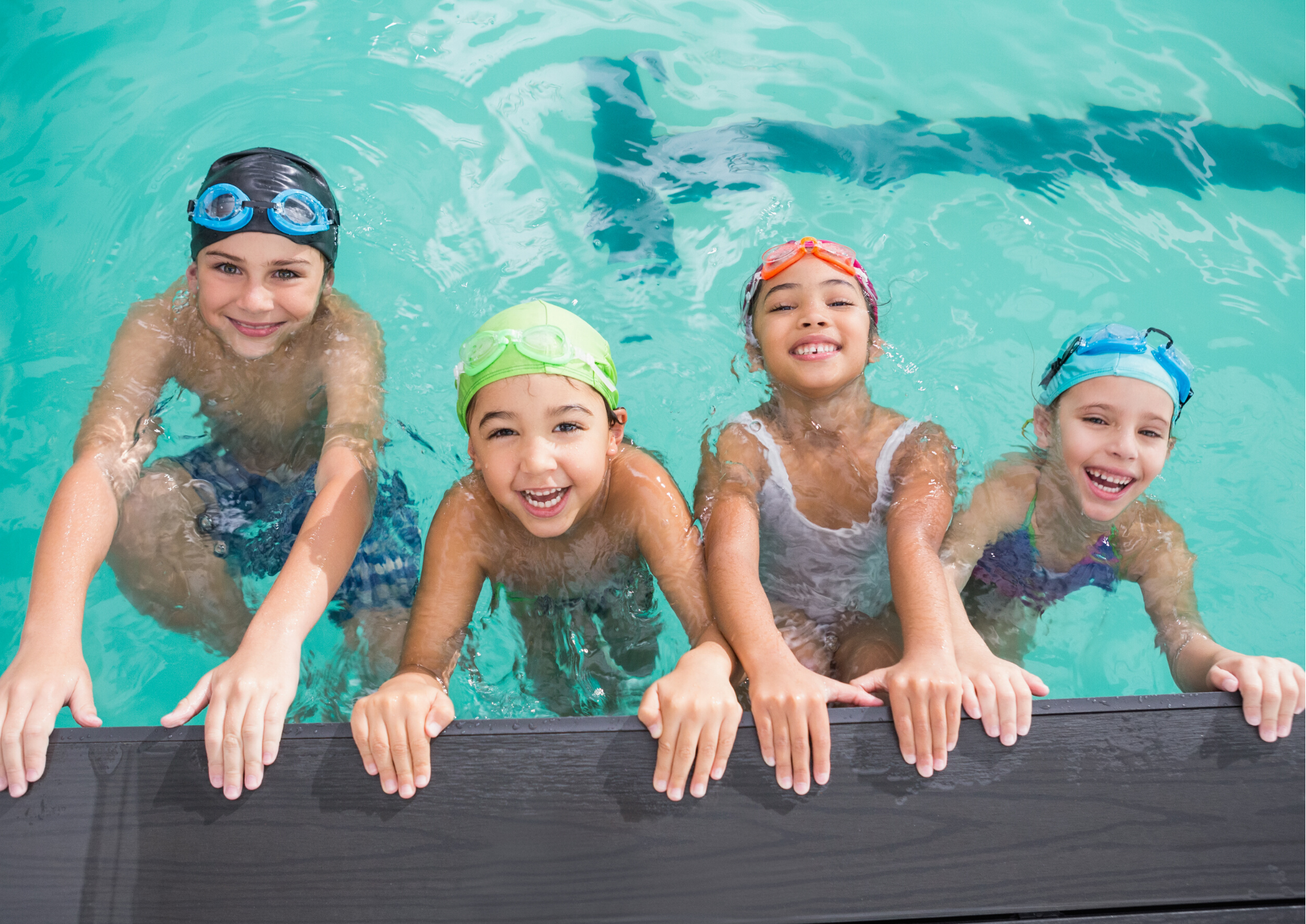 Four cheerful kids in swim gear are smiling and enjoying themselves in a pool, holding onto the edge.