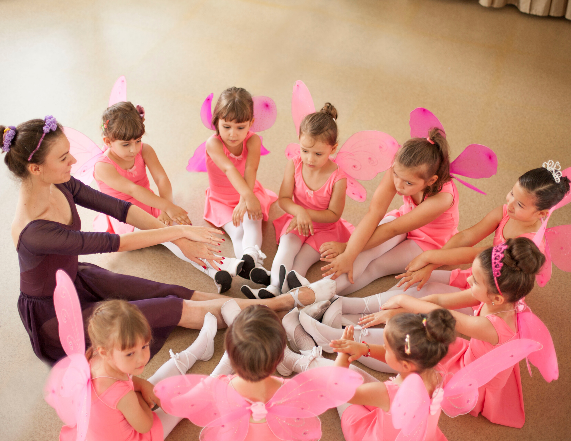 A dance instructor guides a group of young girls in pink dresses and butterfly wings during a ballet class.