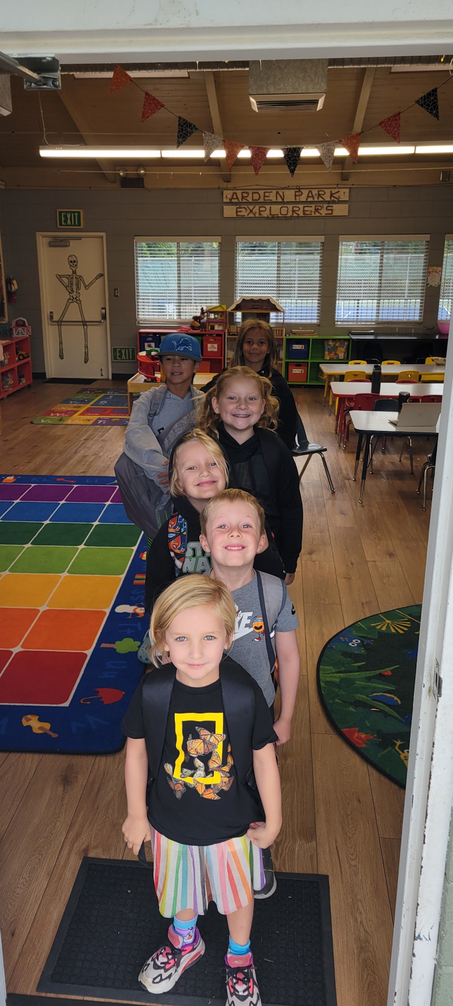 Children posing in a colorful classroom, with educational materials and furnishings. (Note: Image is rotated.)