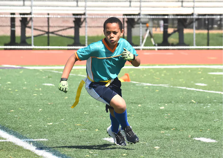 A young boy in a teal and orange sports uniform is running on a football field during practice.