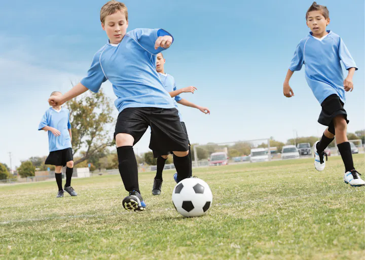 Children in blue jerseys playing soccer on a grassy field under a clear sky.