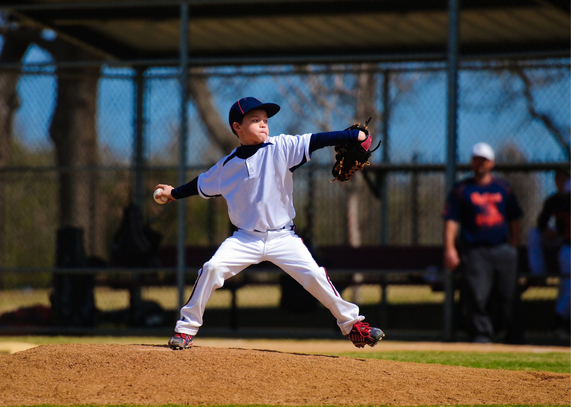 A young baseball player in a white uniform is pitching on the mound with spectators in the background.