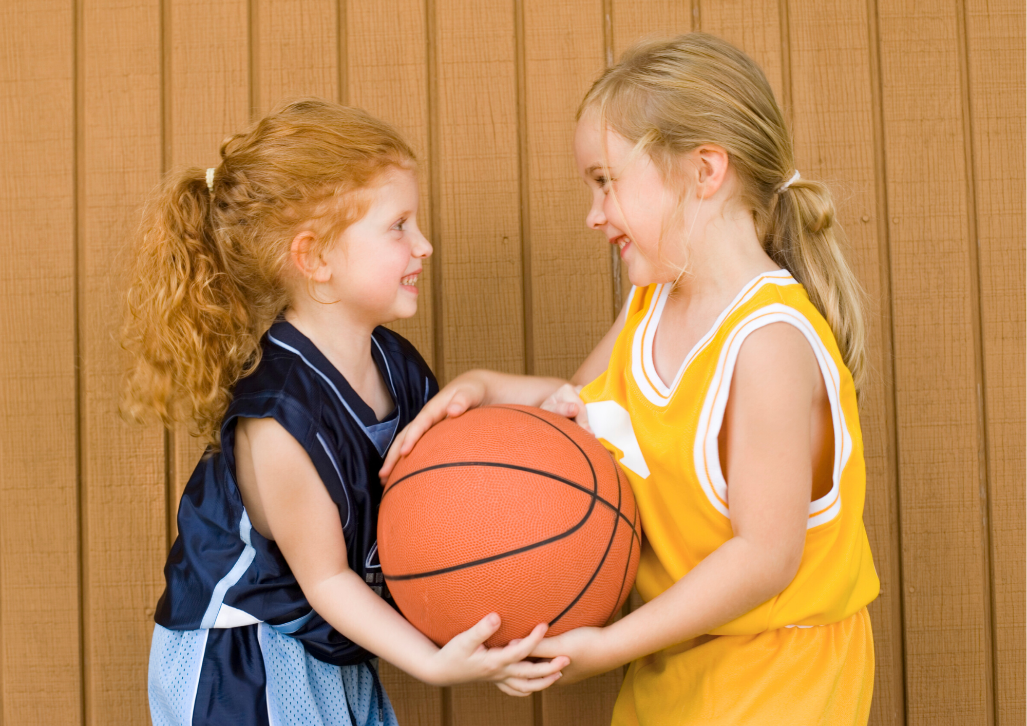 Two young girls, one in a blue basketball jersey and the other in yellow, share a basketball and laugh together.