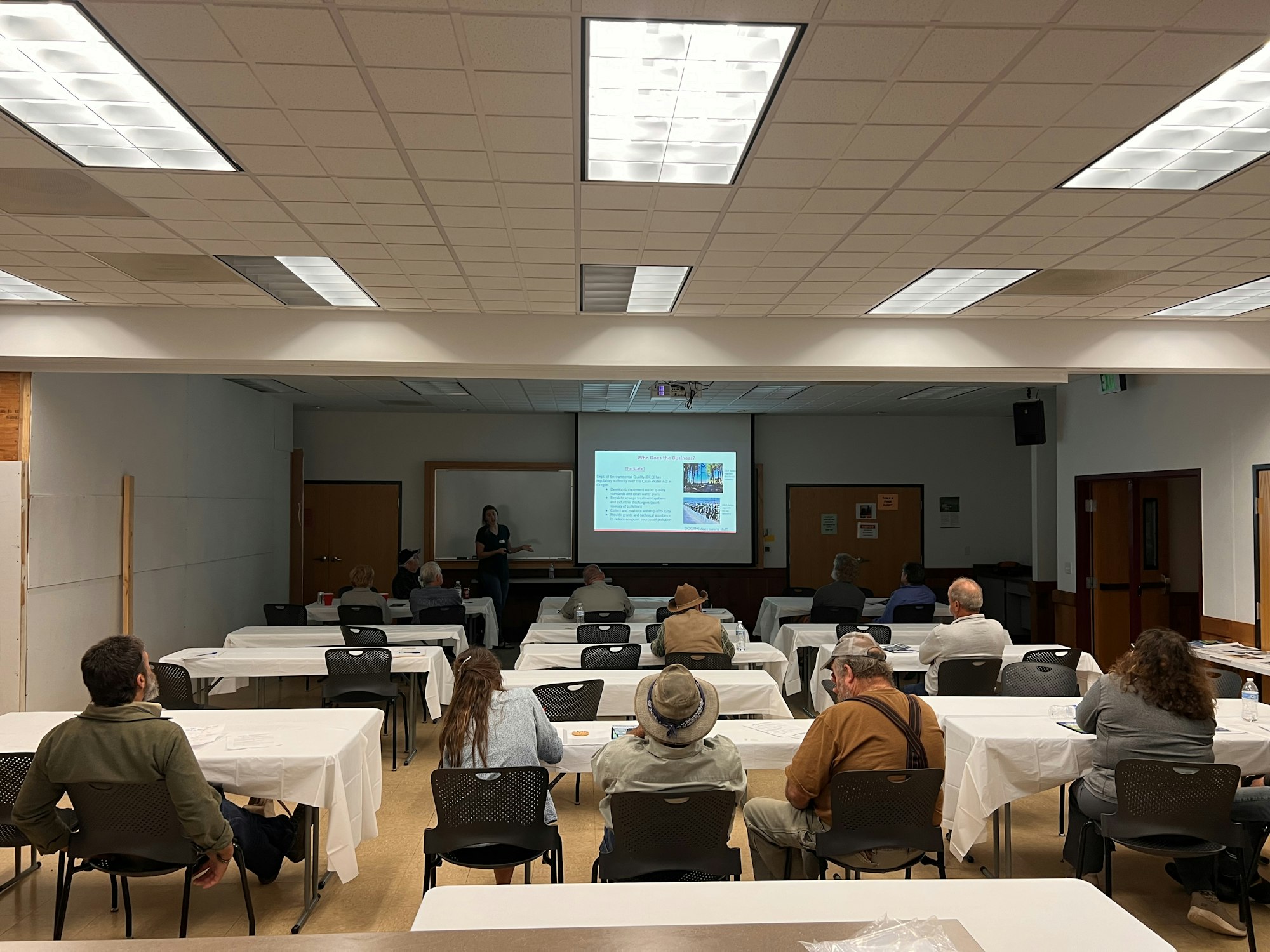 People attending a presentation in a room with tables and a projector screen.