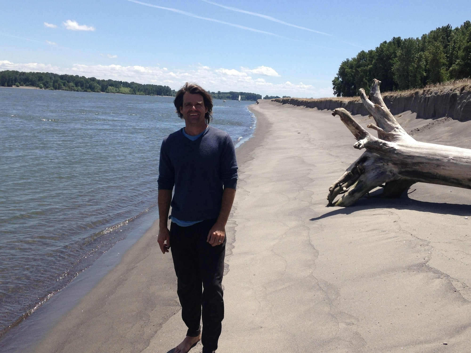 A person standing on a sandy beach near water, with a large driftwood log in the background and trees lining the shore.