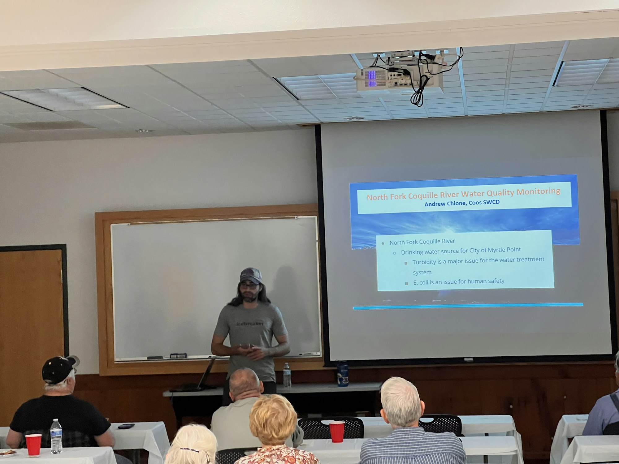 A person presents on North Fork Coquille River water quality monitoring to an audience, highlighting turbidity and E. coli issues.