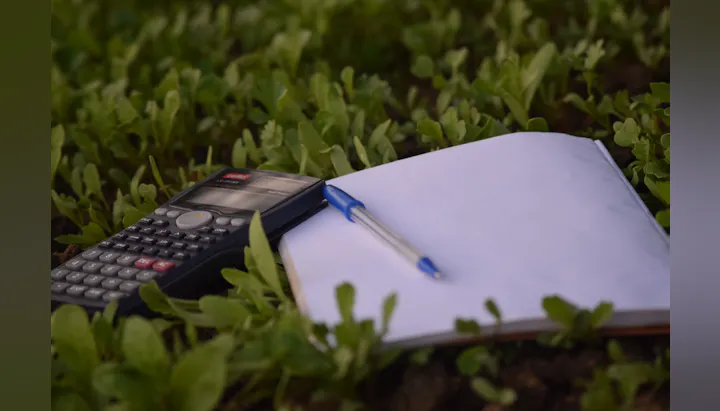 The image features a calculator, a blank notebook, and a pen, set among green plants.