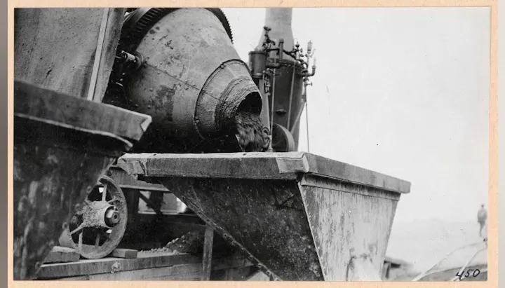The image shows a vintage concrete mixer pouring material into a container, likely for construction or similar purposes.