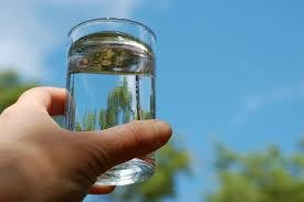 A hand holding a glass of water outdoors, with a background of a blue sky and greenery.
