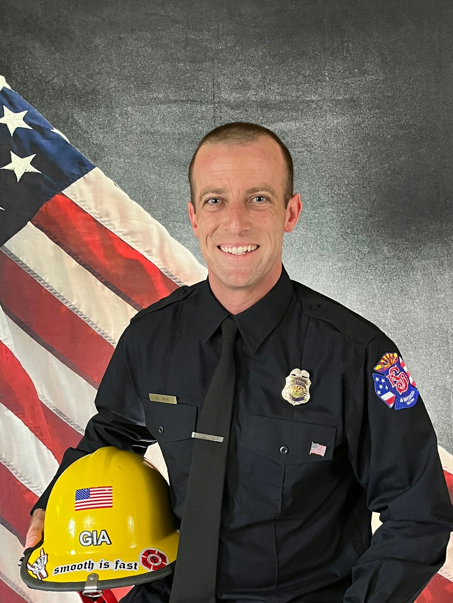 A uniformed individual smiles while holding a yellow helmet with American flags, against a background of the U.S. flag.