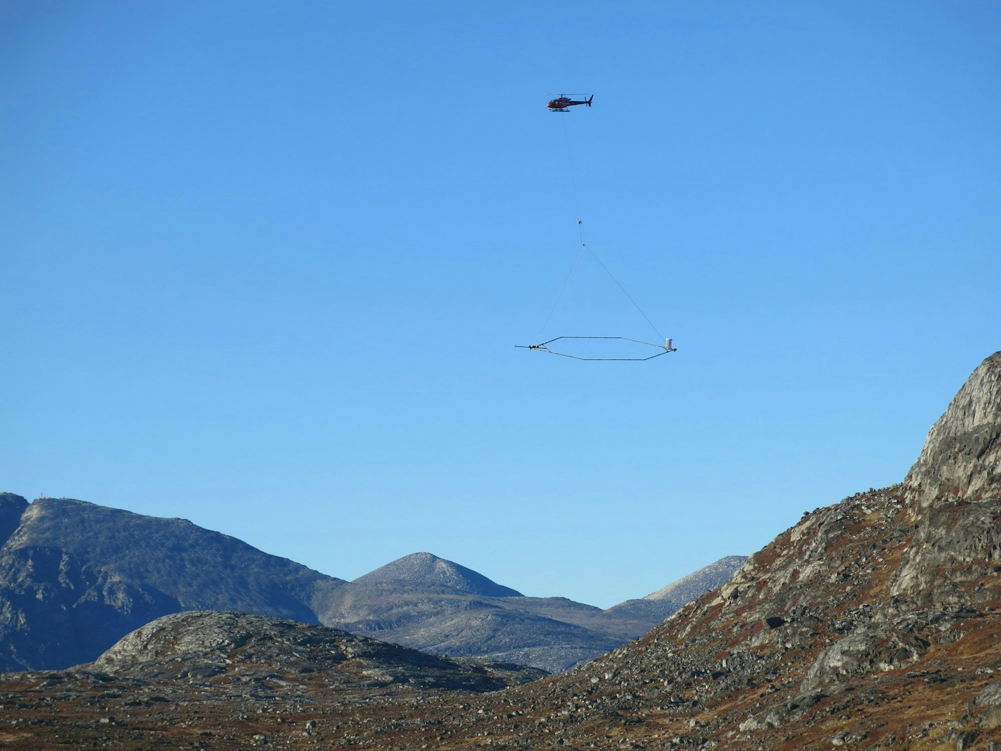Helicopter carrying cargo with a long line over a rugged landscape.