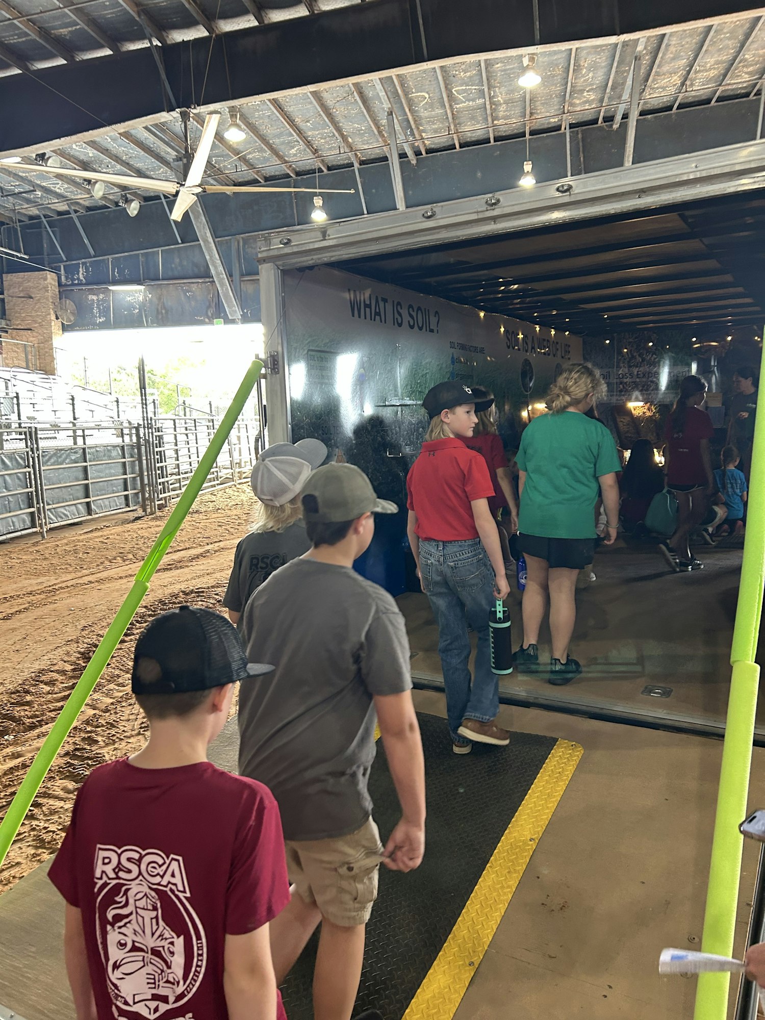 A group of people, mostly children, lined up in a building for an activity or exhibit, with signs and a sandy floor.
