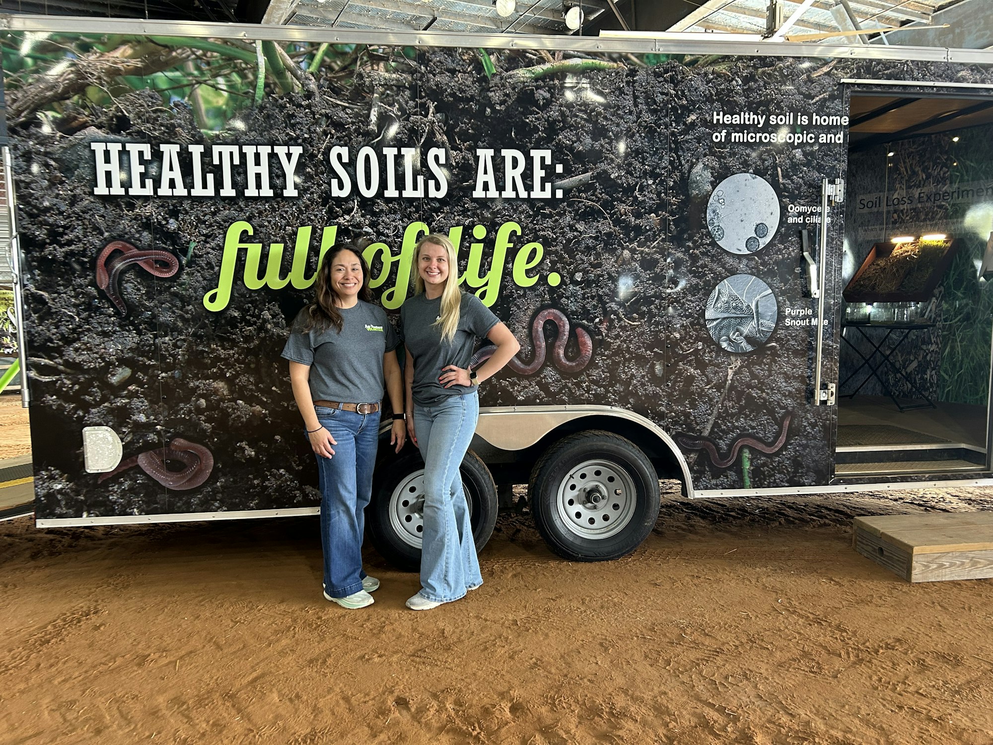 Two women stand by a trailer promoting healthy soils, showcasing the importance of soil diversity and life.
