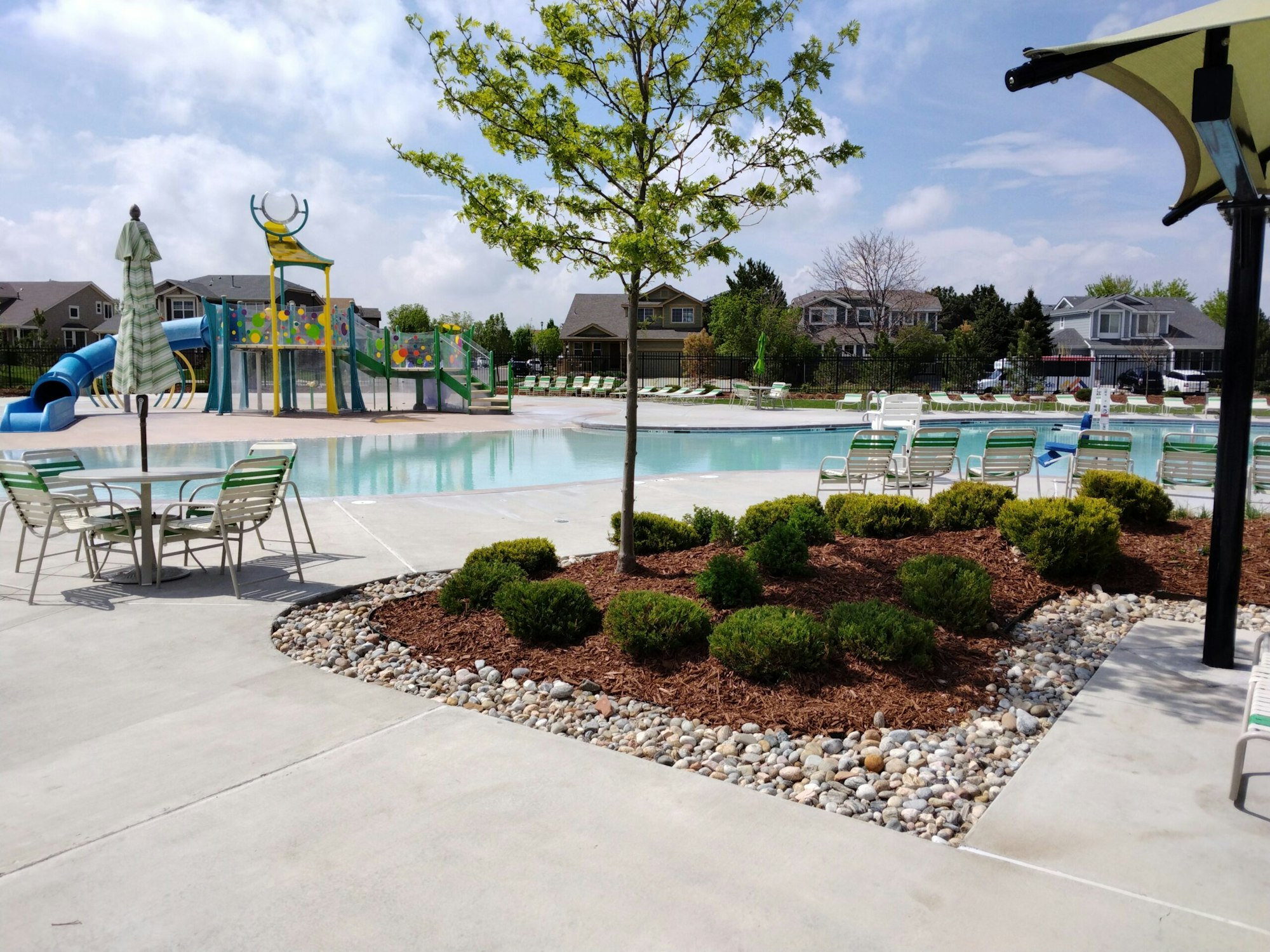 A sunny community pool area with lounge chairs, water slides, and landscaped greenery.