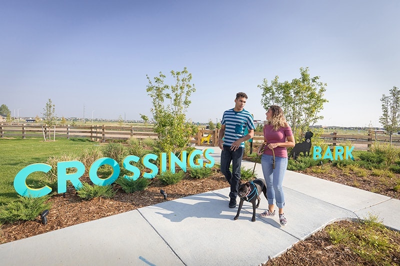 Two people with a dog by "CROSSINGS BARK" sign in a sunny park.