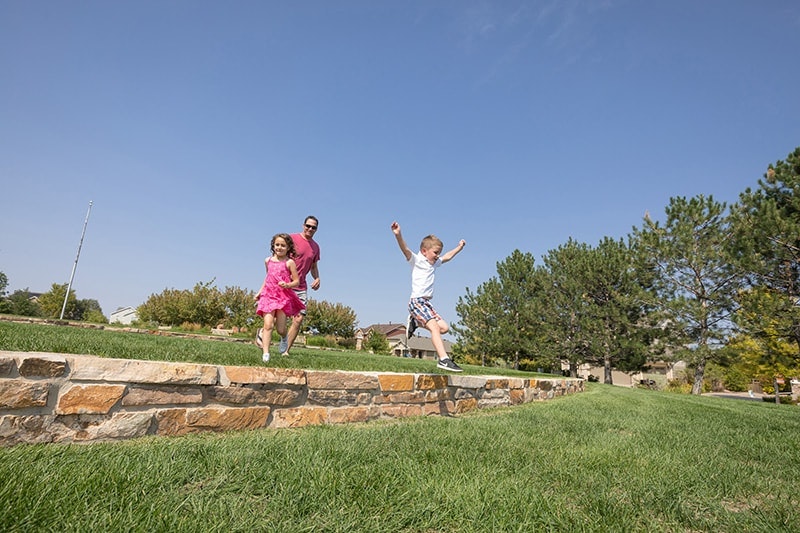 Two kids and an adult are joyfully jumping off a stone wall onto grass under a clear blue sky.
