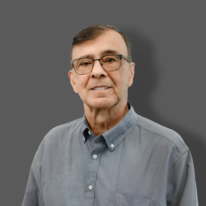 The image features an older man smiling, wearing glasses and a buttoned shirt, set against a grey background.