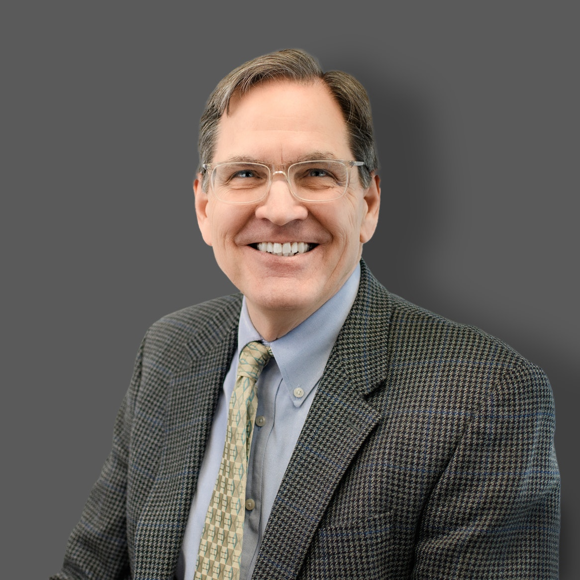 A smiling man with glasses wearing a suit and tie against a grey background.