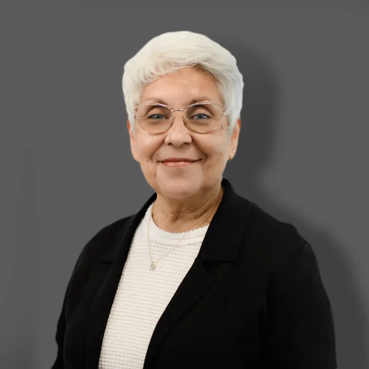 The image shows an older woman with short white hair, wearing glasses and a black sweater, smiling against a gray background.