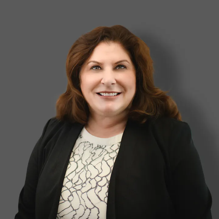 The image features a smiling woman with brown hair, wearing a black blazer over a patterned shirt, set against a gray background.