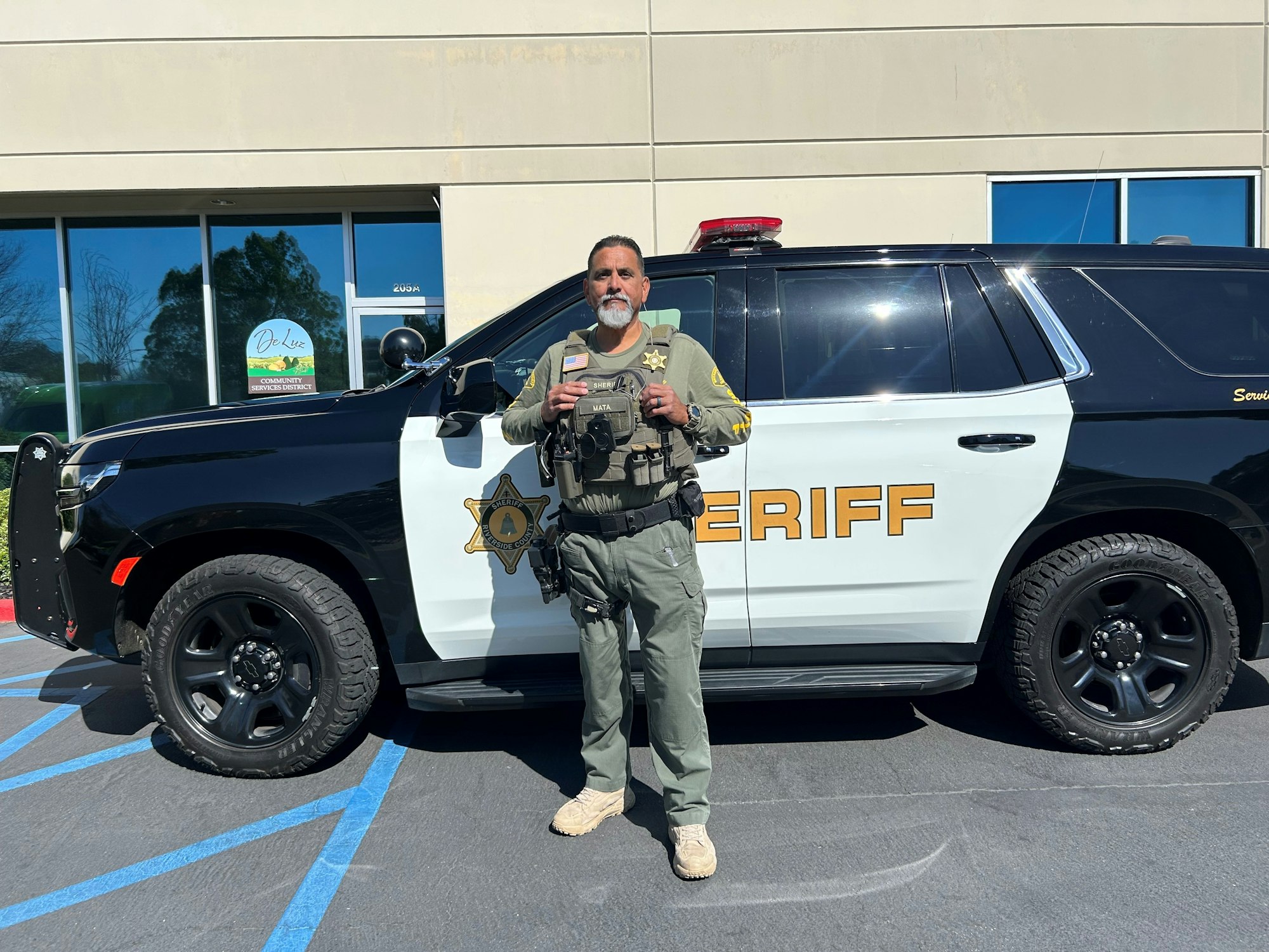 A law enforcement officer stands in front of a sheriff's vehicle outside a building.