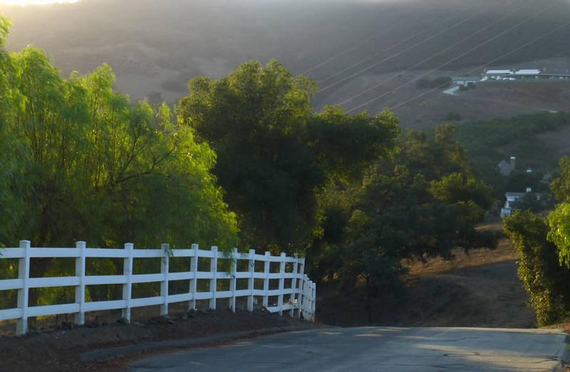 A serene road lined with trees and a white fence, leading toward distant hills and a subtle sunset glow.