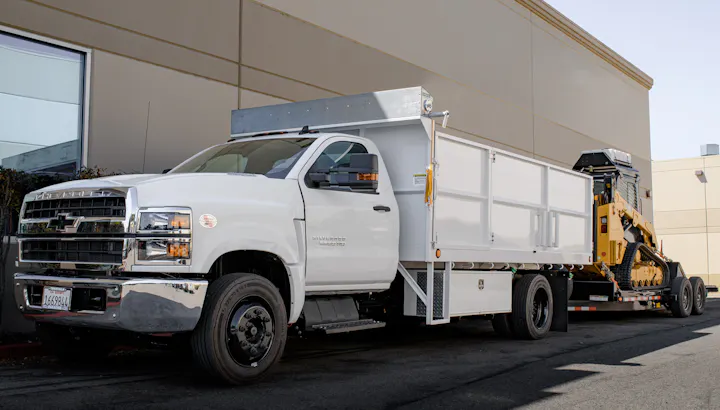 The image shows a white Chevrolet truck with a flatbed trailer carrying construction equipment.