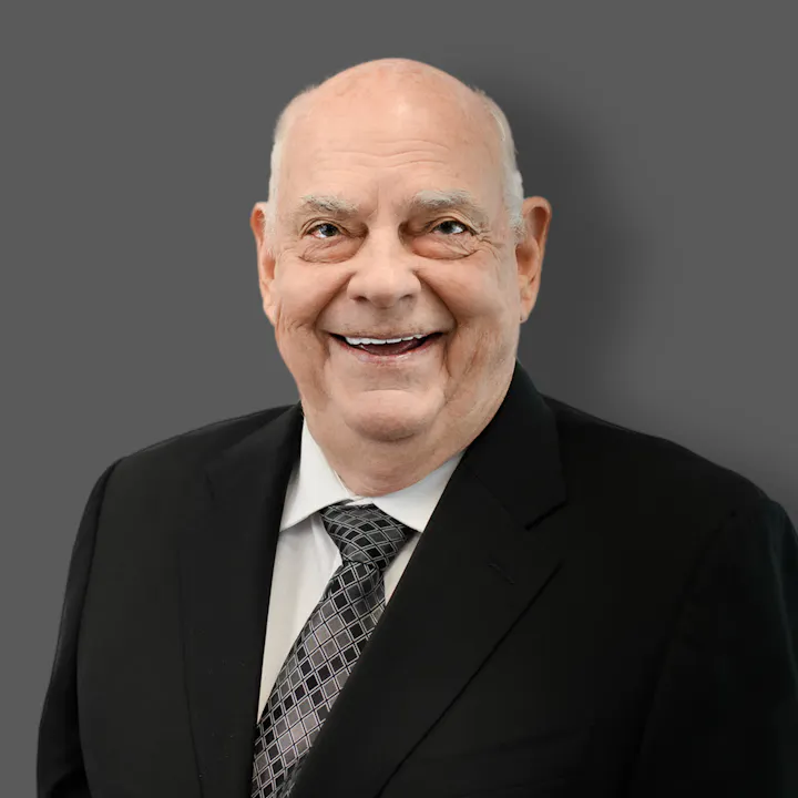 The image features an older man in a black suit and tie, smiling against a gray background.
