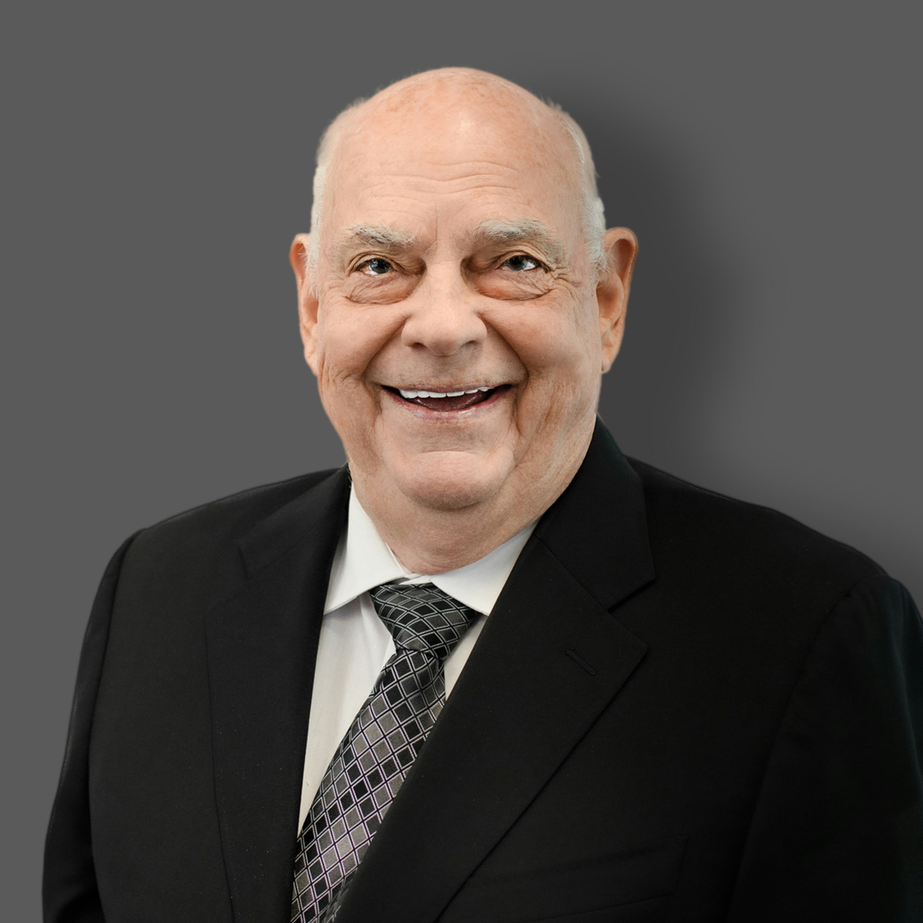 The image features an older man in a black suit and tie, smiling against a gray background.