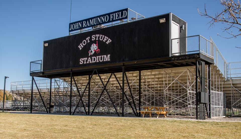 Stadium bleachers with signs for "Hot Stuff Stadium" and "John Ramunno Field," featuring seating structures and a small building.