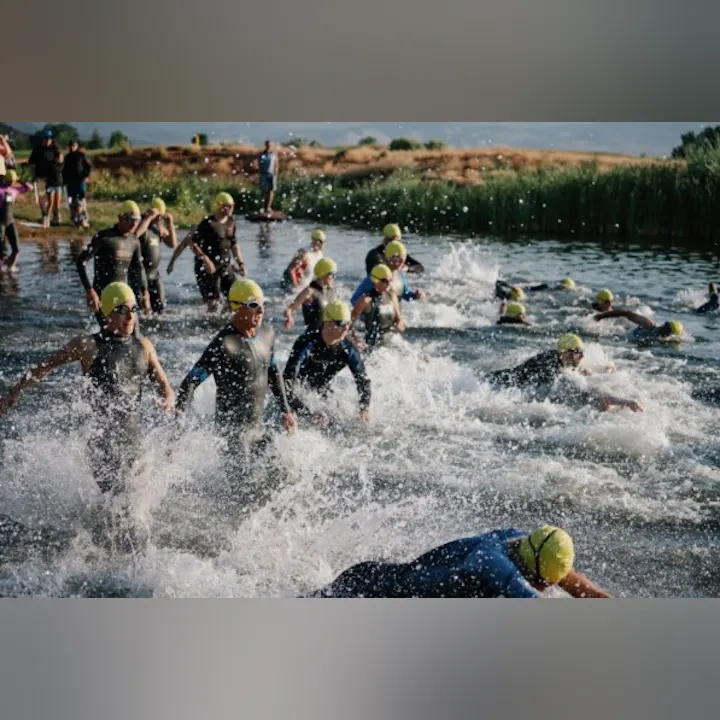 A group of swimmers in wetsuits with yellow caps splash into the water, likely starting a triathlon or swimming competition.