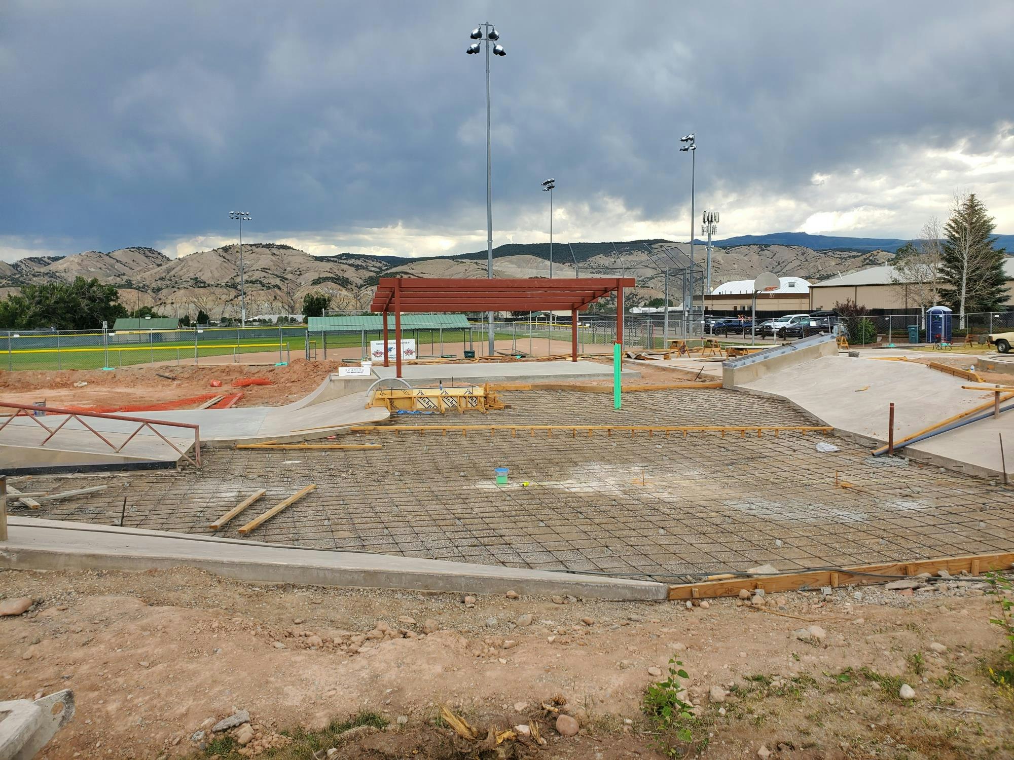 A construction site with metal frameworks, concrete elements, and a red canopy, situated near a sports field under a cloudy sky.