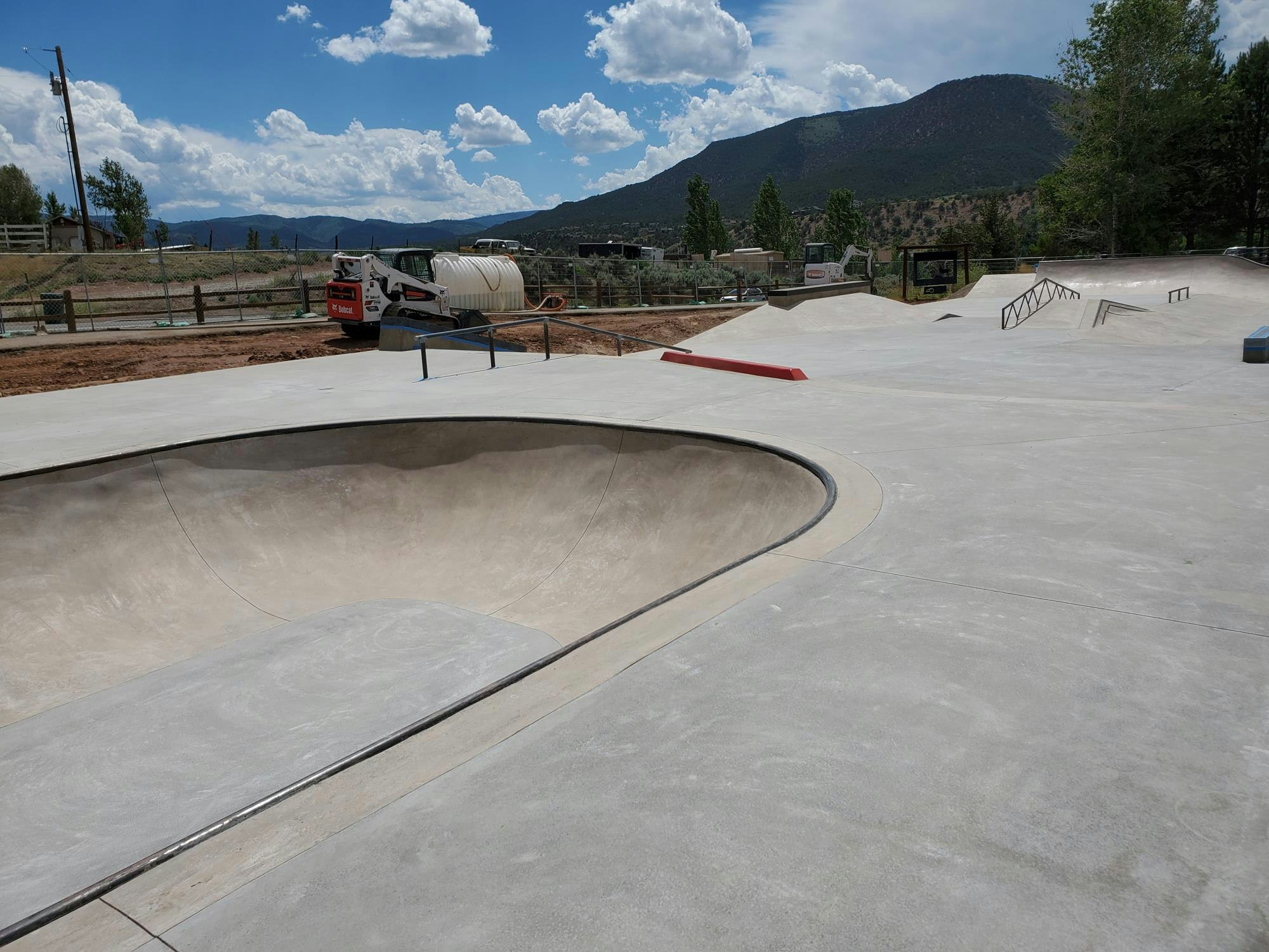 Concrete skatepark with ramps and a bowl, mountains in the background, construction equipment nearby.